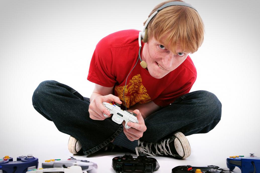 A man sits on the ground in a white room with a headset on, surrounded by game controllers on the floor, as he holds one in both hands