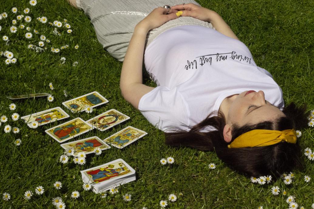 A woman lies on the grass surrounded by daisies with a deck of tarot and a tarot spread laid out next to her