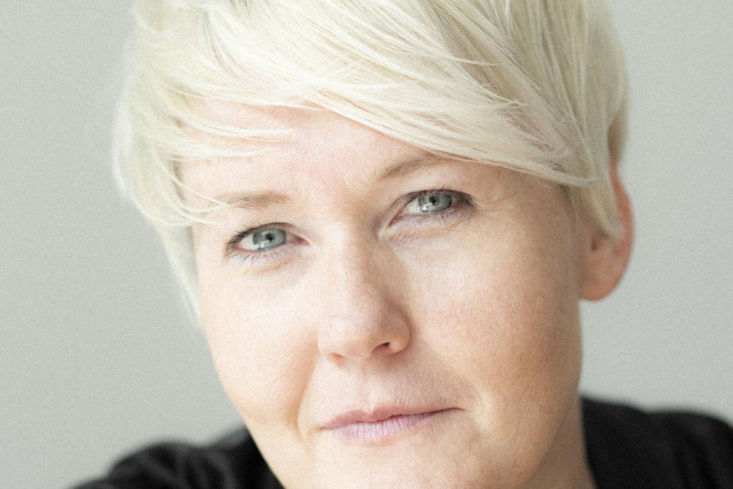 A white woman wearing black clothing with white blond hair looks directly to camera in front of a white background